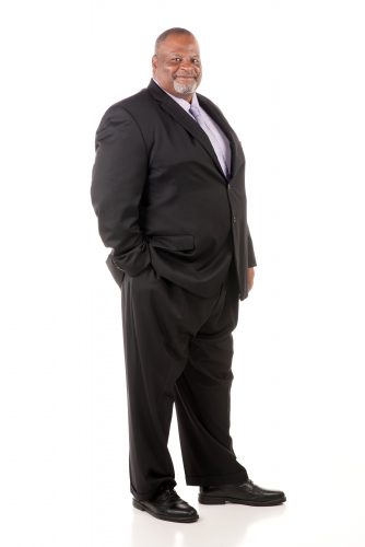 Darron Turner stands on a white floor in front of a white background. He is wearing a black suit with a light purple shirt and tie.