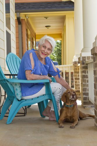 Former American Airlines pilot Beverly Bass with her dog, London, at her Argyle, Texas, home. Bass is sitting in a teal Adirondack chair with a periwinkle shirt, white pants and no shows. The dog is laying on the porch.