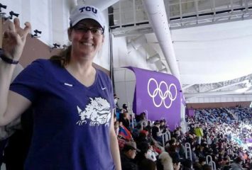 Laura Carmichael Bozeman '92 attended the Men's Figure Skating Finals at the 2018 Winter Olympics at Gangneung Coastal Cluster in South Korea. She is wearing a purple Horned Frog tshirt with her Frog sign up in front of a purple Olympics banner. Photo courtesy of Laura Bozeman