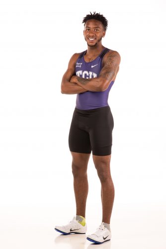 Scotty Newton stands in his TCU uniform (purple top, black shorts, Nike shoes with toes pointed up) against a white background with his arms crossed in front of him, smiling.