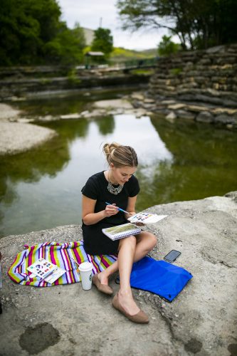 Paris Acklen, an art major, paints on a dry spot in the middle of a waterfall as TCU art students use a variety of media to create art work at Airfield Falls Conservation Park. Photo by Rodger Mallison