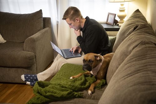 Jonas Kruse sits on a couch with his dog Harley. Jonas is reading his laptop screen. The dog is a boxer.