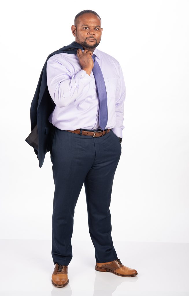 Floyd Wormley stands in front of a white background. He is wearing a blue suit with a light purple shirt and tie. He is holding the suit jacket over his shoulder.