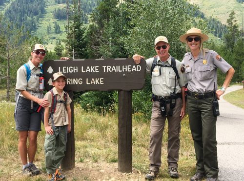 Jess Erwin and a few volunteers of different ages stand in front of a sign directing visitors of Grand Teton National Park to String Lake