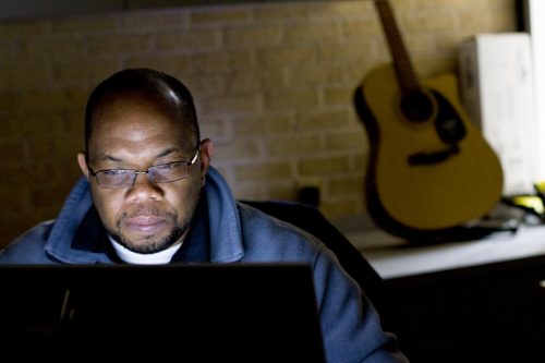 Omar Harvey sits behind his office computer with a guitar in the background