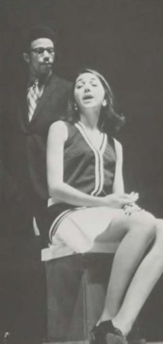 Ronnie Hurdle and a female student perform in a play. The female is sitting and either singing or speaking and Hurdle is standing behind her wearing a suit.