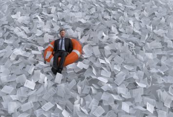 A man wearing a suit sits in a life raft in a sea of paper.