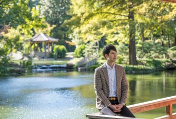 Benjamin Ireland sits on a railing at the Japanese Garden inside the Fort Worth Botanic Garden. He is looking off into the distance and a pond is behind him.