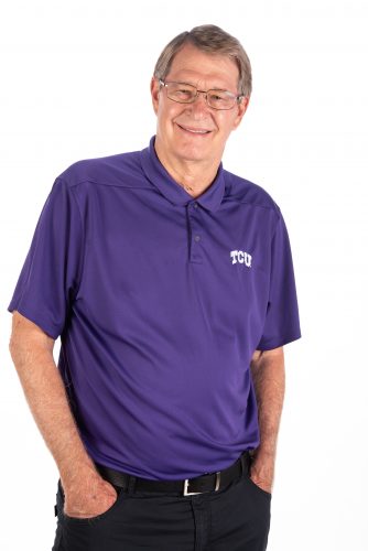 A portrait of head men's golf coach Bill Montigel. He is wearing a purple athletic polo and black slacks with his hands in the pockets.