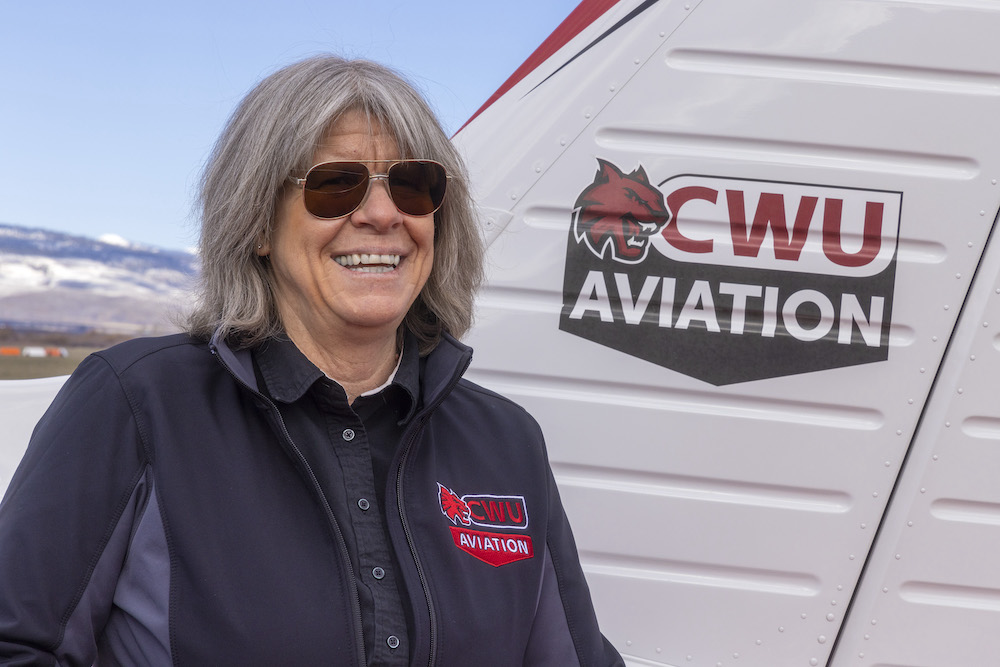 Female pilot in dark sunglasses and a windbreaker smiles in front of a Central Washington University airplane.