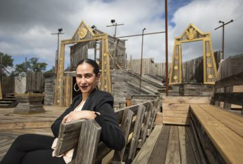 Suited woman sits on a bench with wooden amphitheater set in background.ench, with a wooden theatre set in background.