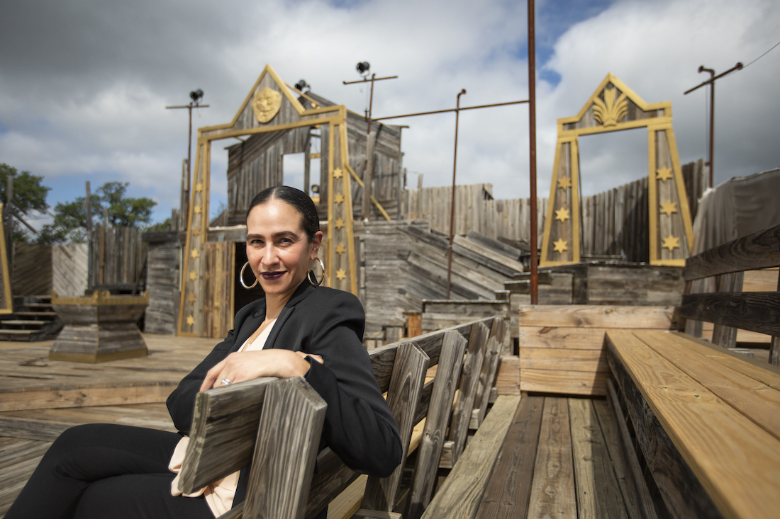 Suited woman sits on a bench with wooden amphitheater set in background.ench, with a wooden theatre set in background.