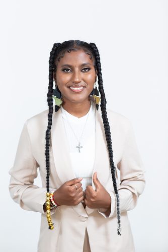 Woman wearing long braids, Africa-shaped earrings, and a white blazer faces the camera.