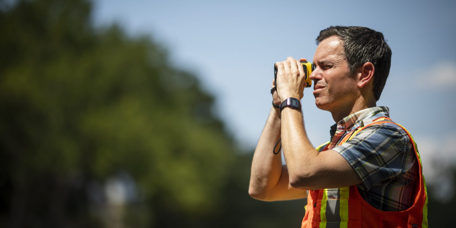 A man in profile, wearing an orange safety vest looks through a device.