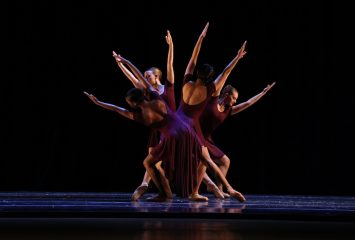 Four dancers in purple, form a starburst-like shape under a spotlight. Their outstretched arms and legs are lit on an otherwise dark stage. .