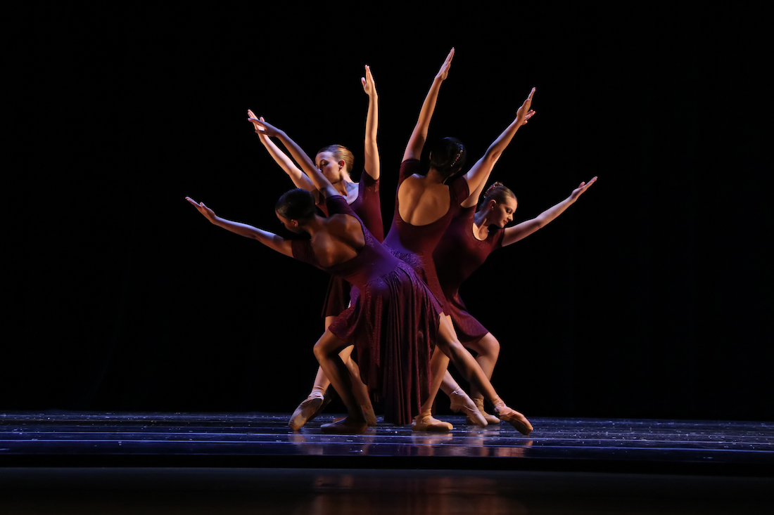 Four dancers in purple, form a starburst-like shape under a spotlight. Their outstretched arms and legs are lit on an otherwise dark stage. .