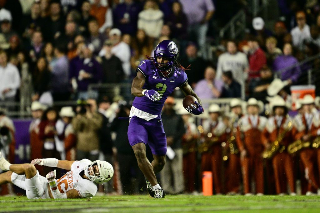 Savion Williams runs in the open field as he cradles a football in his left hand. In the background, Texas defensive back Michael Taaffe is sprawled across the grass, having failed to tackle Williams.