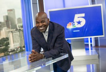 Sportscaster Newy Scruggs leans against a desk on the set of NBC 5, the station's logo stretched across a blue screen over Scruggs' left shoulder. Scruggs smiles toward the camera, his elbows resting on the desk and his hands in front of him.