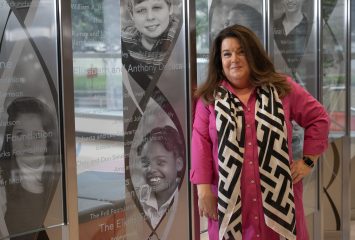 A photograph of Angela Synek, director of advancement at the Duncan Neurological Research Institute at Texas Children's Hospital in Houston. Synek, pictured in a pink garment with a black and white patterned scarf hanging from her neck, stands for a portrait photograph on the ground floor of the Jan and Dan Duncan Neurological Research Institute Building in Houston, Texas.