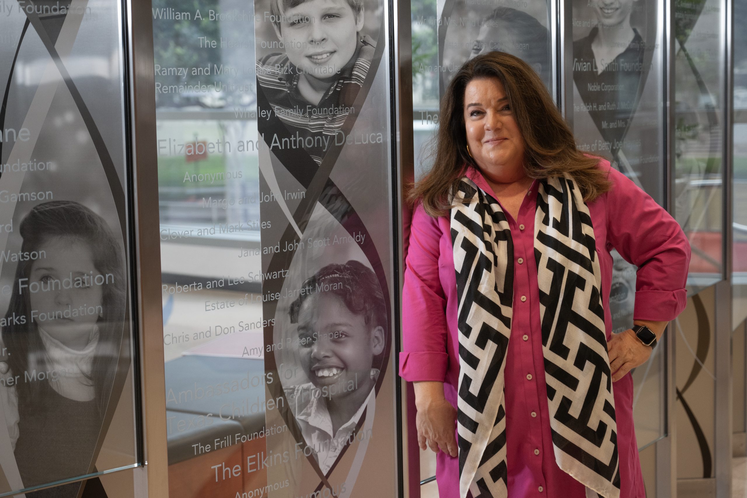 A photograph of Angela Synek, director of advancement at the Duncan Neurological Research Institute at Texas Children's Hospital in Houston. Synek, pictured in a pink garment with a black and white patterned scarf hanging from her neck, stands for a portrait photograph on the ground floor of the Jan and Dan Duncan Neurological Research Institute Building in Houston, Texas.