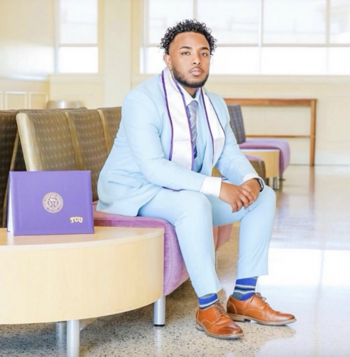 Image of Mohamed Ade Mohamed wearing a powder blue suit and brown dress shoes. The Class of 2022 TCU graduate sits with a purple diploma placed on the coffee table to his right. Ade Mohamed has his hands crossed above his knees as he looks toward the camera.