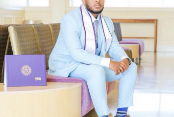 Image of Mohamed Ade Mohamed wearing a powder blue suit and brown dress shoes. The Class of 2022 TCU graduate sits with a purple diploma placed on the coffee table to his right. Ade Mohamed has his hands crossed above his knees as he looks toward the camera.