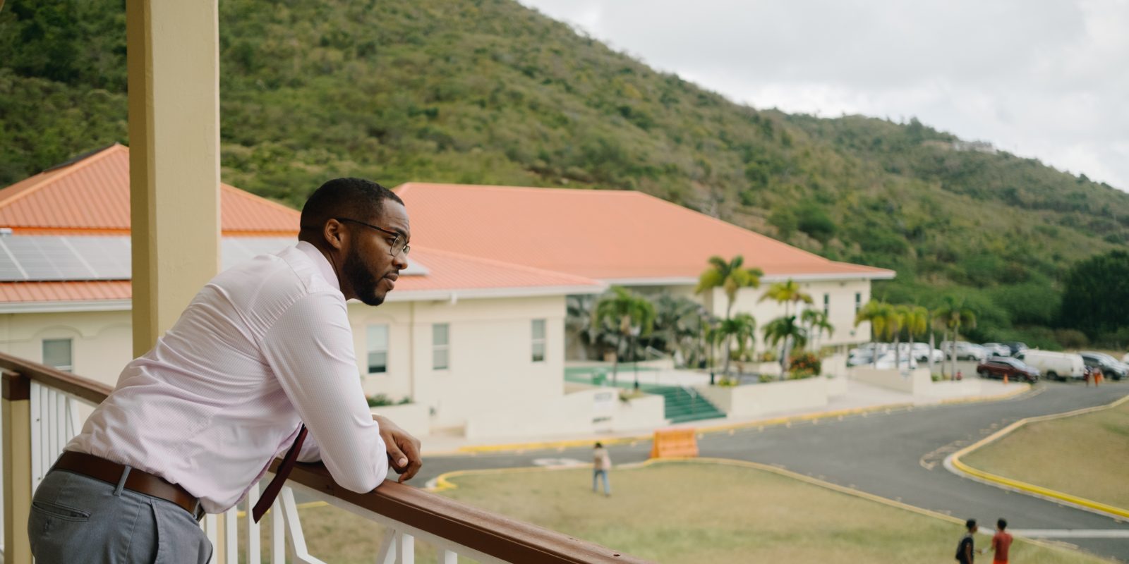 Richard Georges leans forward at a three-quarters angle away from the camera with his forearms resting on a balcony railing. A red-roofed, tan building fills the left side of the background, while a vegetation-rich mountain rises above the building toward the top-left corner of the frame.