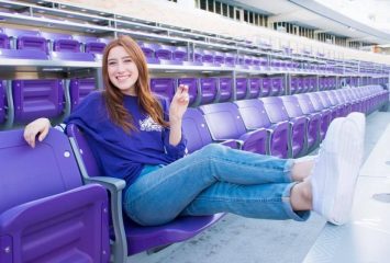 A photograph of TCU alum Erin Dianis, dressed in a purple TCU Horned Frogs t-shirt and jeans, seated in a purple arena chair. Dianis makes the TCU hand symbol with her left hand as she kicks her feet up in front of her.