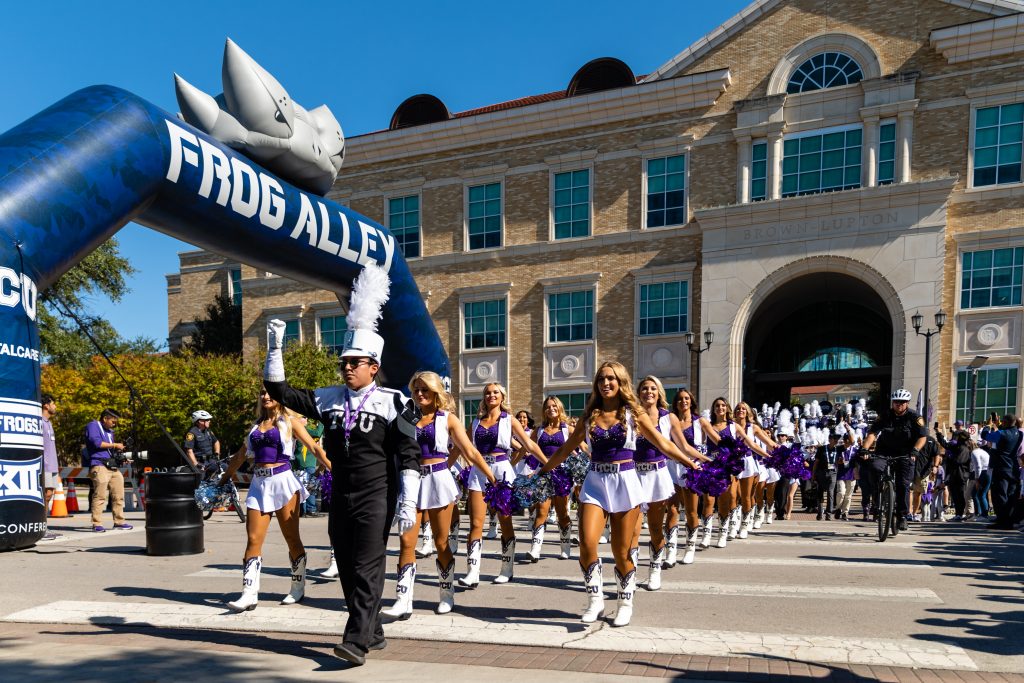 A 2021 photograph of Frog Alley before a Big 12 football game against the Baylor Bears. Led by a color guard, TCU cheerleaders emerge from the Brown-Lupton University Union, leading fans into the stadium.