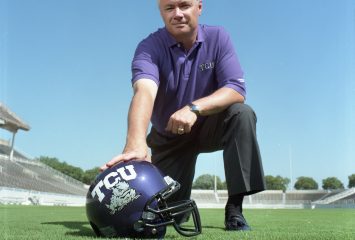 Head coach Dennis Franchione kneels at center frame, with his left forearm resting on his left knee and his right hand on the top of a TCU football helmet. Franchione is pictured on a football field as he grins toward the camera on a sunny, cloudless day.