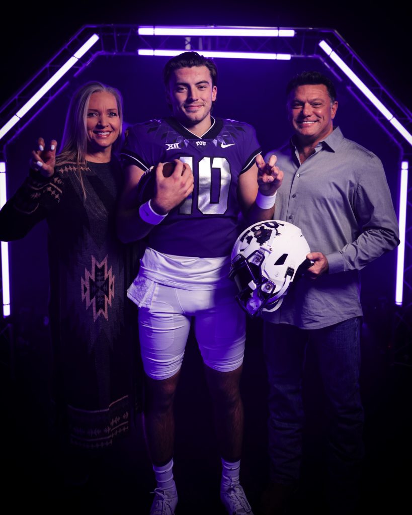 Ken Seals, who is wearing a purple TCU football jersey, flashes the "go frogs" hand sign with his left hand as he holds a football with his right. He's standing alongside his father to his left and his mother to his right.