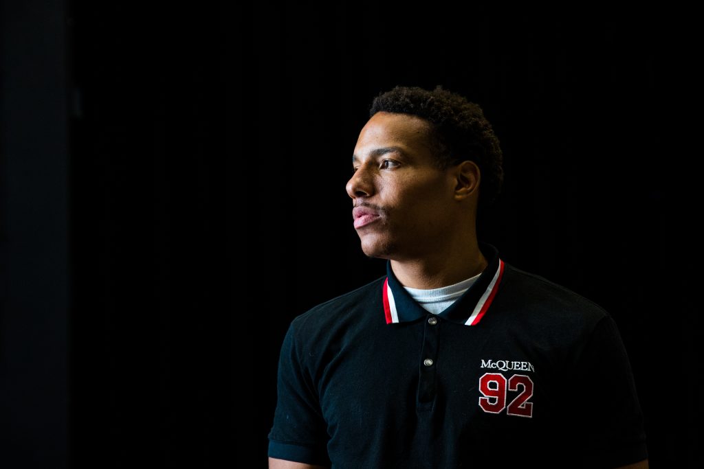 Desmond Bane, wearing a black polo shirt with red and white text embroidery, looks to his right. A light shines upon Bane's face. He's pictured against a black background. Bane is involved with youth advocacy through a camp he started for kids facing difficulties with coordination.