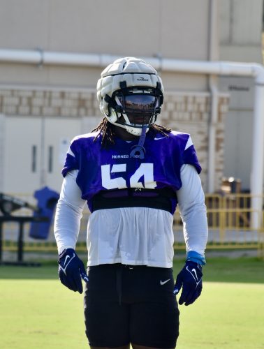 Photograph of TCU student-athlete NaNa Osafo-Mensah standing on the practice field during fall camp. Osafo-Mensah is wearing a visored helmet. His purple mouthguard hangs down from the bottom rung of his face mask. He is wearing a purple practice jersey bearing the number 54. He's wearing a white long-sleeve undershirt, purple gloves and black Nike-branded football pants.