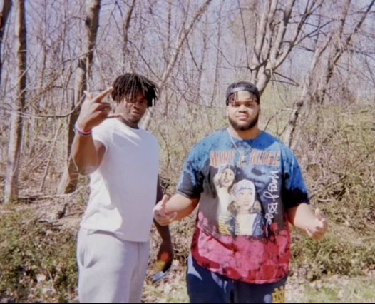 A photograph of friends Nana Osafo-Mensah and Jacob Lacey. The two stand in front of a wooded area. Osafo-Mensah, wearing a white t-shirt, holds his right hand toward the camera at shoulder height. Lacey, wearing a black baseball cap and a blue, black and red tie-dye shirt, holds up each of his hands at waist height, with his palms facing upward.