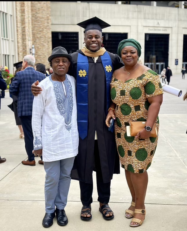 A photograph of NaNa Osafo-Mensah standing between his parents Kwaku (left) and Joyce (right). Osafo-Mensah wraps his hands around his parents' shoulders and smiles toward the camera. He is wearing his Notre Dame graduation garb, complete with a black robe and graduation cap as well as a blue graduation stole embroidered with gold Notre Dame "ND" logos on either side.