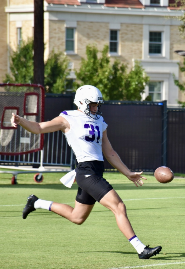 Photograph of TCU punter Ethan Craw, in his white helmet, white practice uniform and black shorts, kicking a football at fall camp.