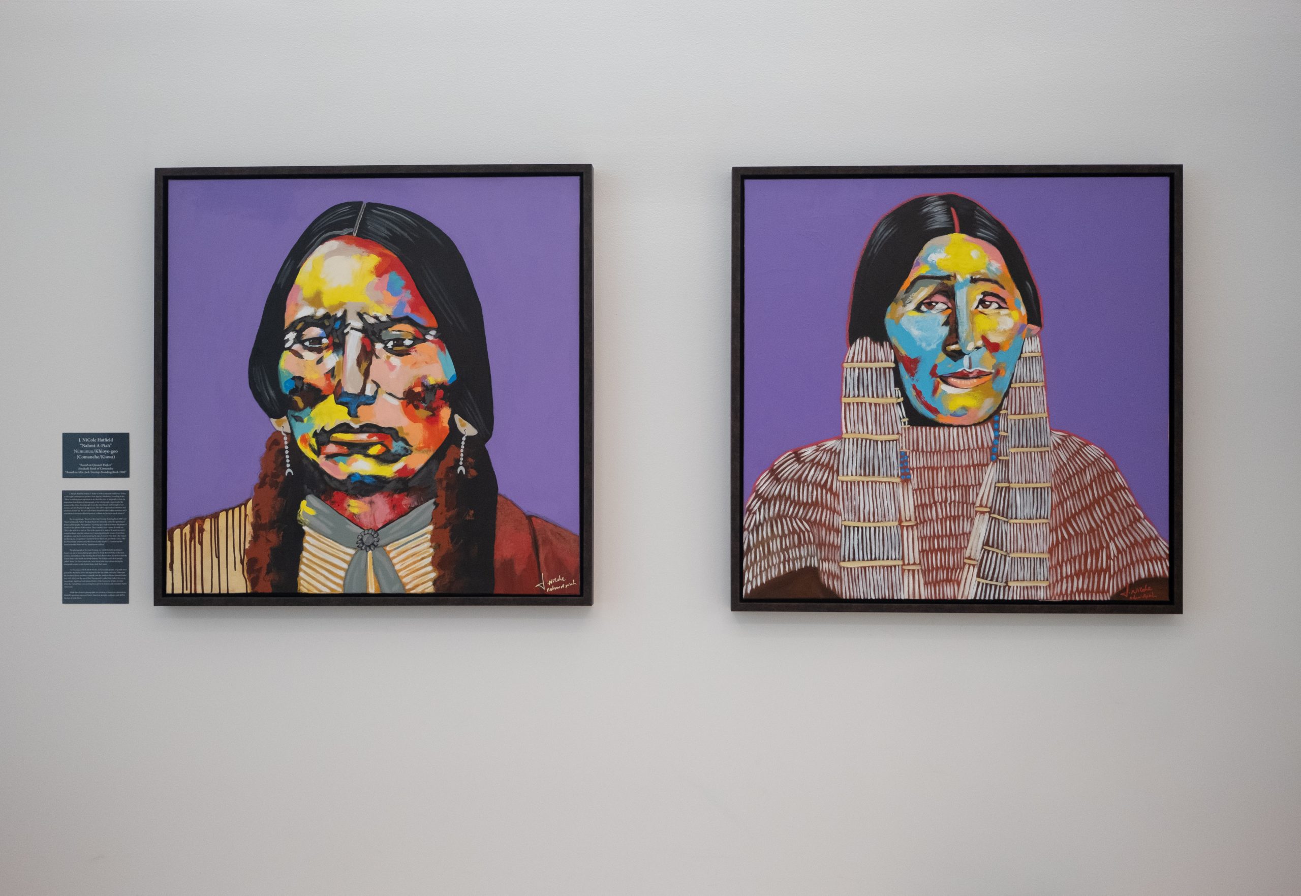 Image of two paintings by J. NiCole Hatfield (Nahmi-A-Piah) hung on one of the Harrison building's conference room walls. Described as "kaleidoscopic paintings," both works contain plain lavender backgrounds and figures composed of vibrant-colored paint.