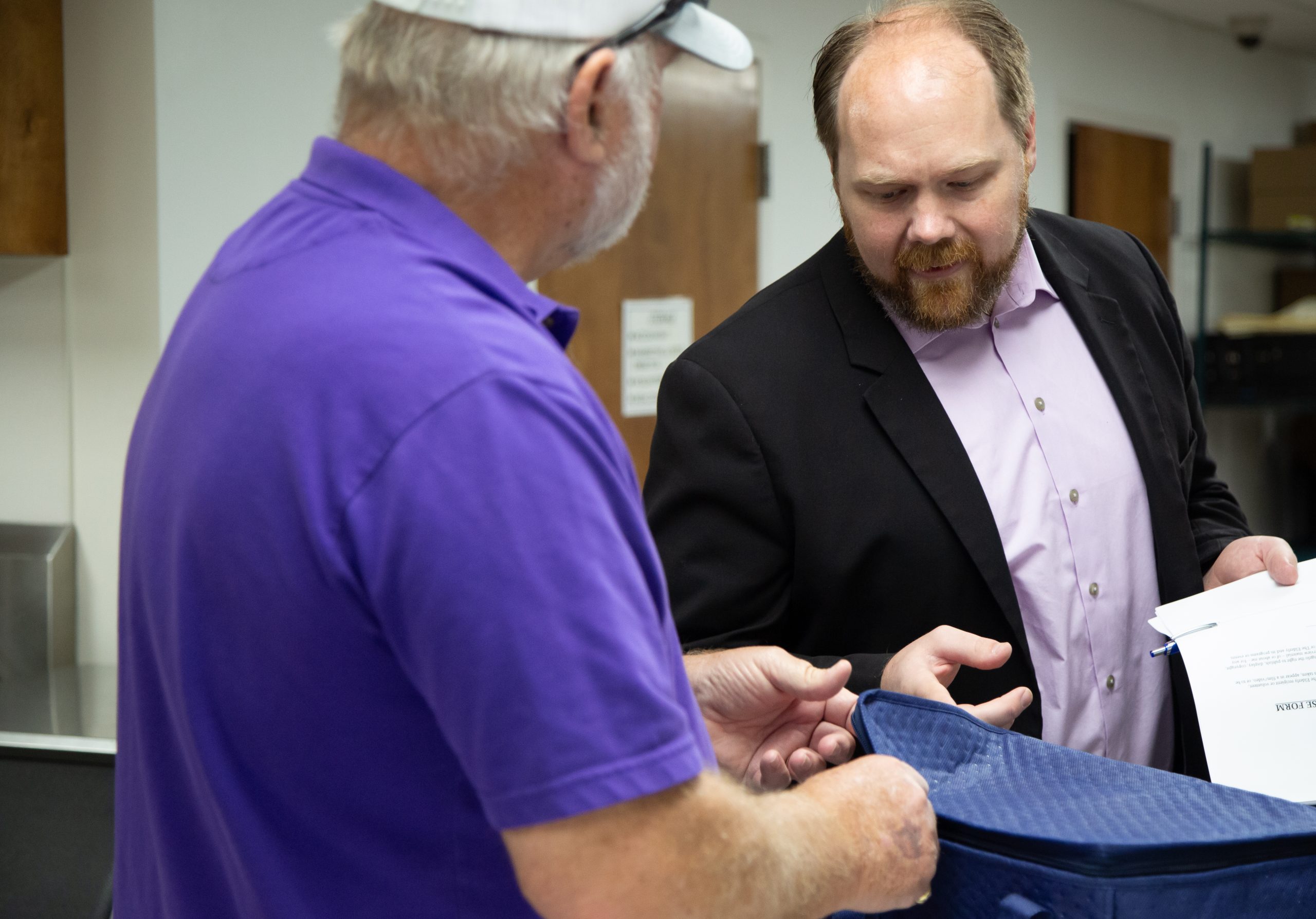 Dason Tucker points toward a navy blue cooler bag as a white-bearded man, wearing a purple shirt and white hat, and standing at a third-quarters pivot away from the camera, unzips the cooler.