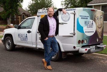 Dason Tucker, standing with his right hand in his jean pocket and his left arm leaning against a white Meals for the Elderly truck, smiles toward the camera.