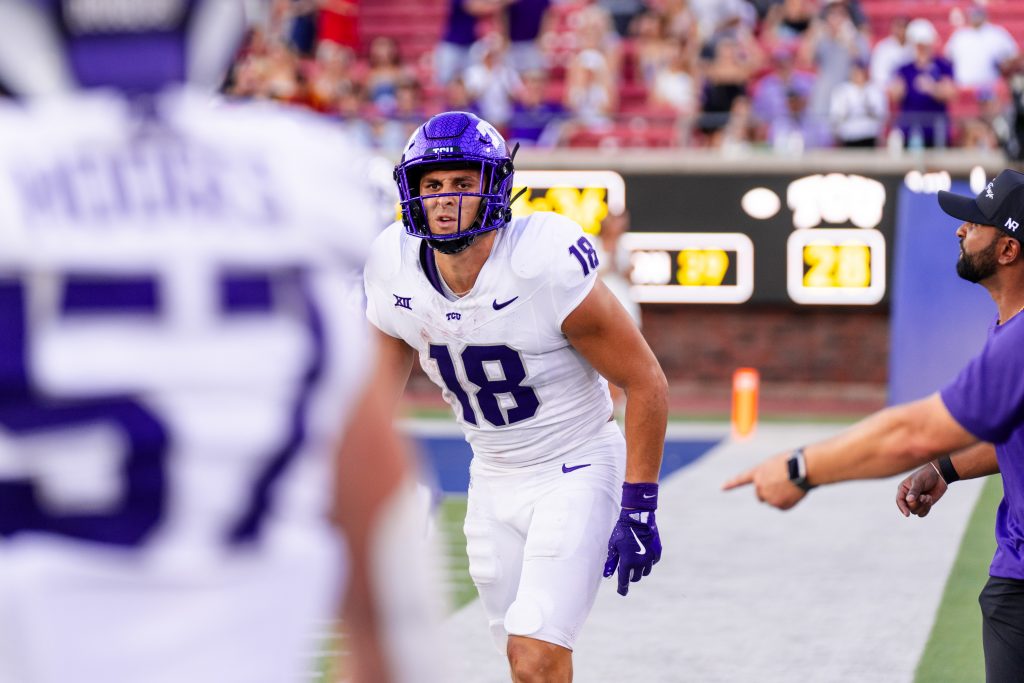 Photograph of Jack Bech walking back toward the TCU sideline. The back of linebacker Johnny Hodges fills the left-hand side of the image. Both student-athletes are wearing TCU football's white road uniforms with purple helmets.