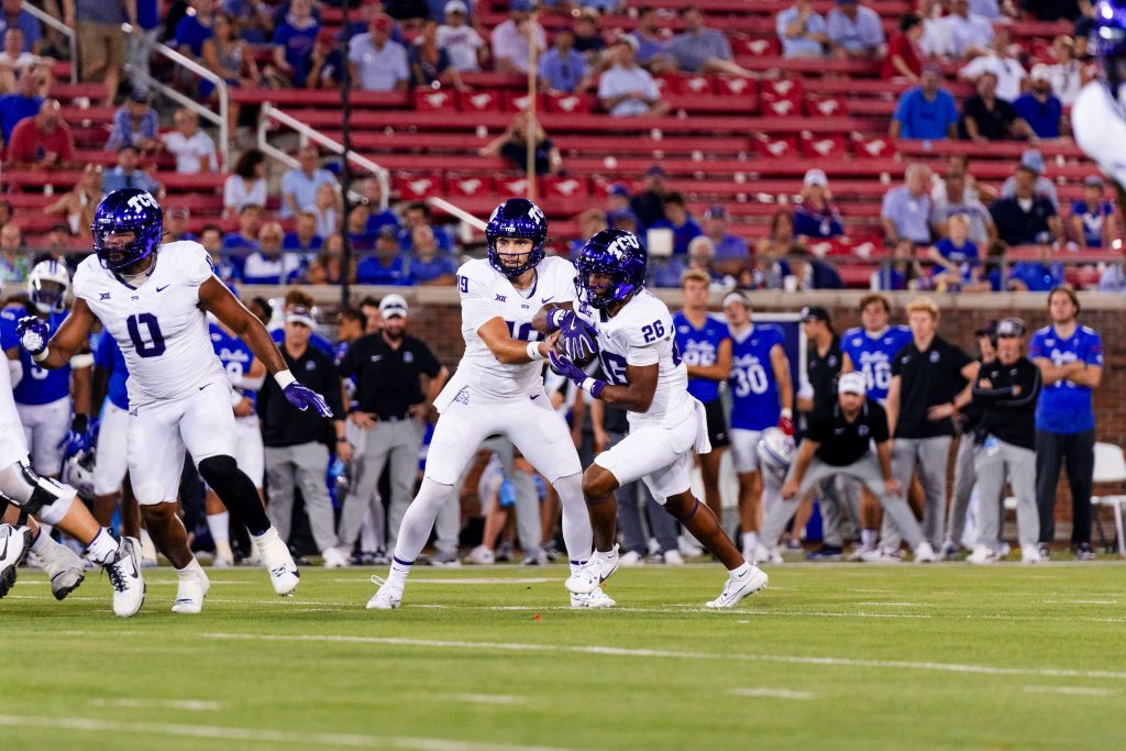 Photograph of quarterback Ken Seals handing the football to running back Jeremy Payne. Both student-athletes are wearing TCU's white road football uniforms with purple helmets. In the background, SMU coaches and players watch from the sideline.