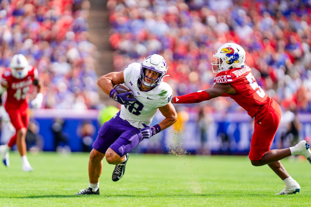 Photograph of TCU wide receiver Jack Bech running by a Kansas defender during a college football game at Arrowhead Stadium. Bech is wearing a white football helmet, white jersey and purple pants. He bends away from the Kansas player's outstretched hand as he works his way upfield. 