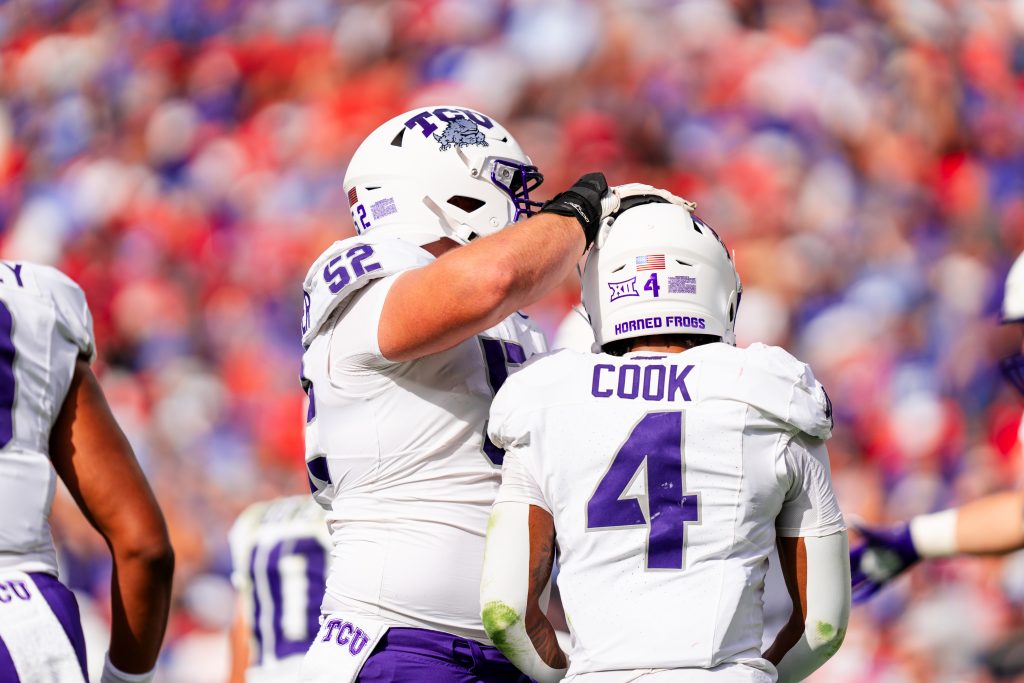 Photograph of offensive lineman James Brockermeyer tapping running back Cam Cook on the helmet. Both players are facing away from the camera and are wearing white TCU football helmets and white jerseys with purple lettering.