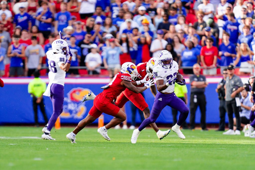 Photograph of TCU running back Jeremy Payne carrying the ball during the Horned Frogs' Sept. 28 game against Kansas at Arrowhead Stadium. Payne, wearing a white TCU football helmet, a white jersey embroidered with the number 26, purple pants and white cleats, carries the ball in his left hand as he runs by two Jayhawk defenders. The Kansas players, in white helmets, red uniforms and pants and white cleats, extend toward Payne in an effort to tackle him. Wide receiver Eric McAlister is in the image's background, as are fans in the stands behind him.