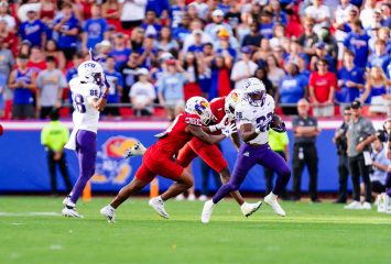 Photograph of TCU running back Jeremy Payne carrying the ball during the Horned Frogs' Sept. 28 game against Kansas at Arrowhead Stadium. Payne, wearing a white TCU football helmet, a white jersey embroidered with the number 26, purple pants and white cleats, carries the ball in his left hand as he runs by two Jayhawk defenders. The Kansas players, in white helmets, red uniforms and pants and white cleats, extend toward Payne in an effort to tackle him. Wide receiver Eric McAlister is in the image's background, as are fans in the stands behind him.