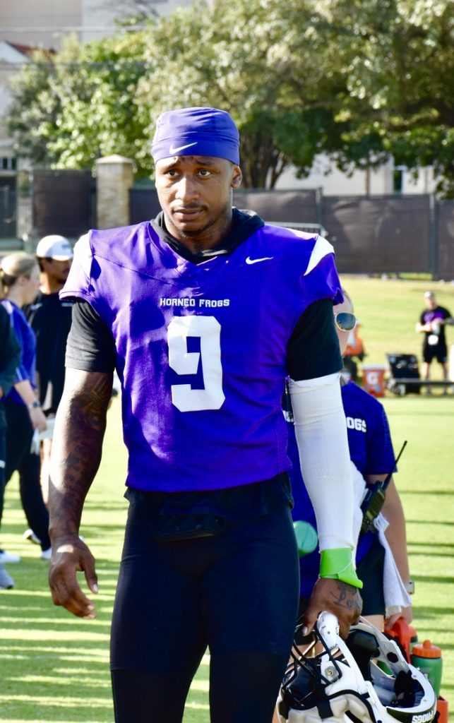 Photograph of Marcel Brooks during TCU Football's 2024 fall camp. Brooks is wearing a purple Nike skullcap, a purple Horned Frogs practice jersey and a black short-sleeve undershirt with a white sleeve on his left forearm. Brooks has lime green trainer's tape on his left wrist and is holding his football helmet in his left hand as he looks toward the camera. 