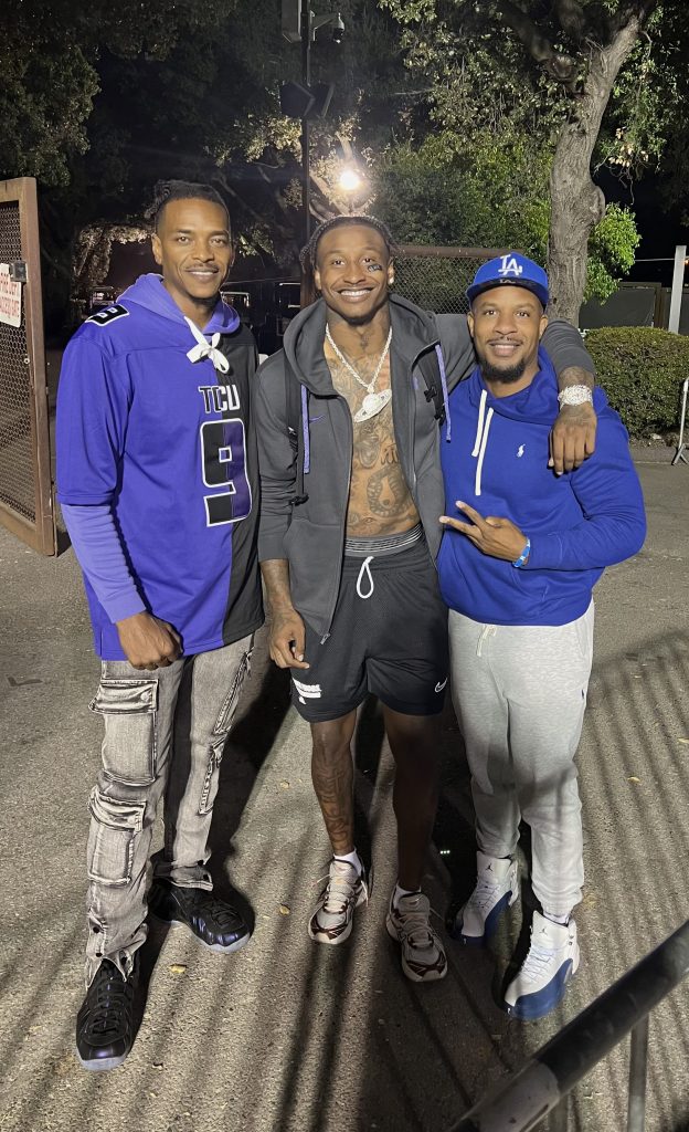 Photograph of Marcus Brooks with his father, William, and his uncle, Don. All three are standing and smiling toward the camera. Marcus stands in the center with his left arm around the shoulder of his uncle, Don, who is to Marcus's left. Marcus wears a grey sweatshirt with black shorts and white and black sneakers. Don, wearing a blue Los Angeles Dodgers hat, blue sweatshirt, grey sweatpants and white and blue sneakers, holds up a peace sign with his left hand. William stands to Marcus's right. He's wearing a black, purple and white number 9 TCU football jersey, grey jeans and black sneakers.