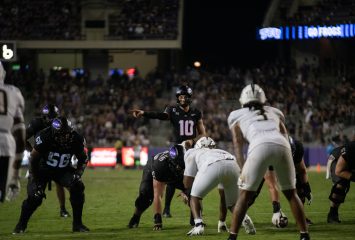 Photograph of TCU quarterback Josh Hoover making pre-snap adjustments during a Sept. 14 game against UCF. Hoover, wearing TCU's all-black football uniform, puts to his right as the offensive line remains in their stance in front of him. UCF defenders dot the foreground of the photograph.