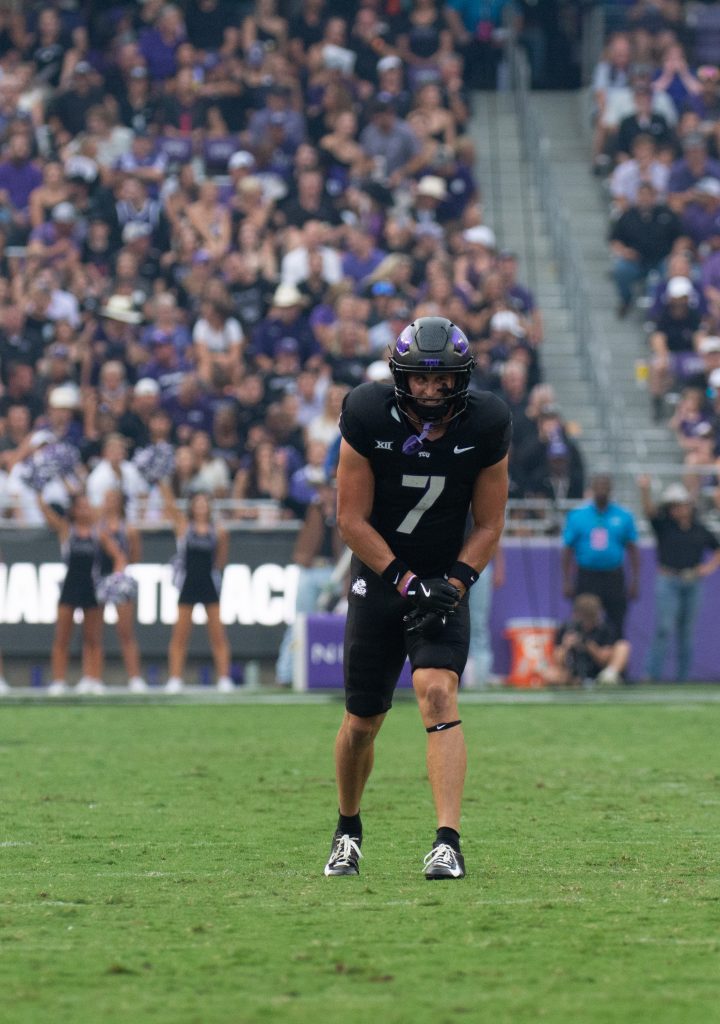 Photograph of TCU wide receiver JP Richardson, standing in his pre-snap stance during TCU Football's Sept. 14 game against UCF. Richardson is leaning forward on his left knee, with both hands placed on his left thigh. Grass fills the lower third of the photo's background. The high background of the photo shows fans sitting in the stands.