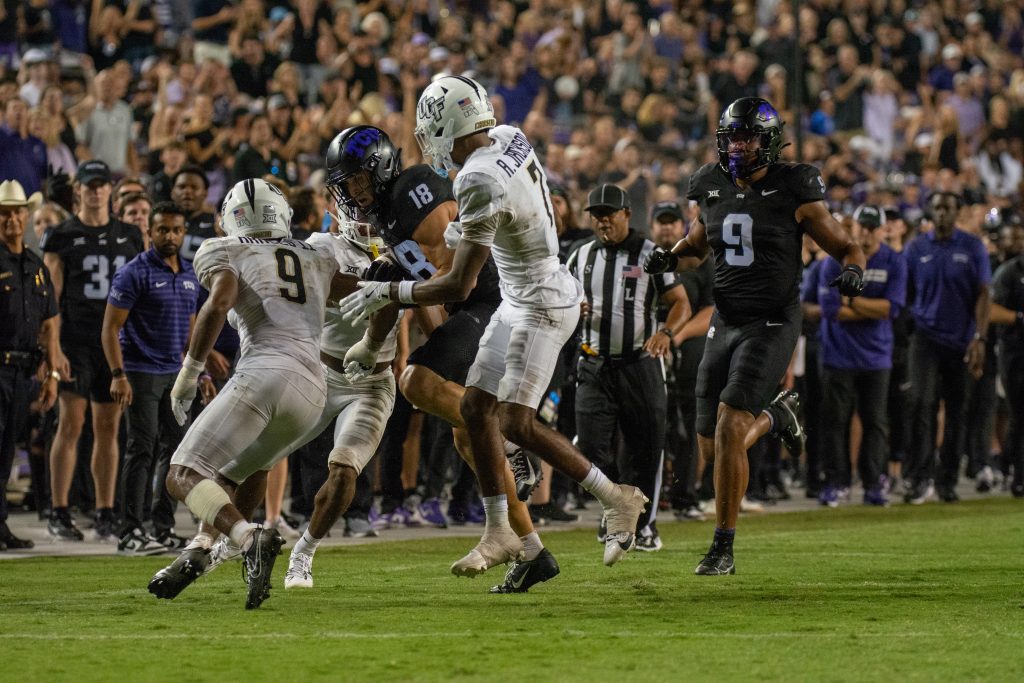 Photograph of Jack Bech encircled by three UCF defenders. Bech, wearing number 18 and clad in an all-black TCU uniform, cradles the ball in his right hand as he braces for contact. Bech's teammate, Drake Dabney, is pictured in the background.